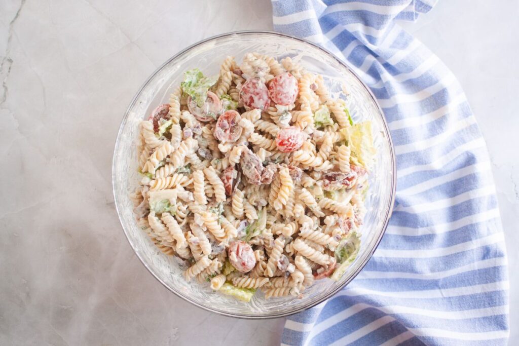 Overhead shot of a glass bowl filled with pasta salad, featuring spiral pasta, tomatoes, bacon, lettuce, and creamy dressing. A blue striped cloth is visible on the side.