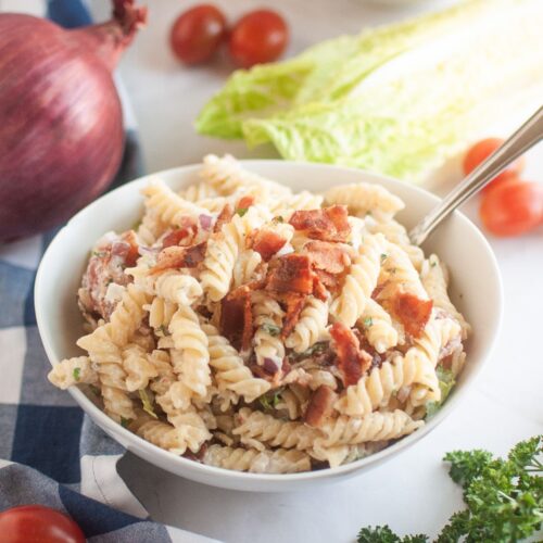 A white bowl filled with pasta salad, featuring spiral pasta, creamy dressing, pieces of bacon, and other visible ingredients, is shown with a spoon. Red onions, lettuce, cherry tomatoes, and parsley are in the background.