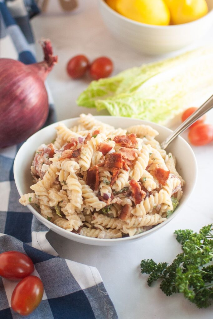 A white bowl filled with pasta salad, featuring spiral pasta, creamy dressing, pieces of bacon, and other visible ingredients, is shown with a spoon. Red onions, lettuce, cherry tomatoes, and parsley are in the background.
