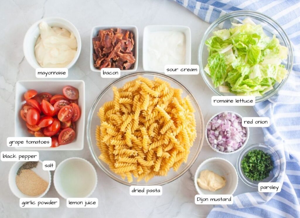 Overhead shot of various ingredients for a pasta salad, including cooked spiral pasta in a glass bowl, bowls of bacon, lettuce, red onion, parsley, tomatoes, and dressing components, arranged on a marble surface.