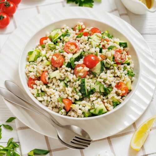 A bowl of barley salad with cherry tomatoes, cucumber, and herbs, served with a fork and spoon on a plate, surrounded by dressing, parsley, lemon, and condiments.