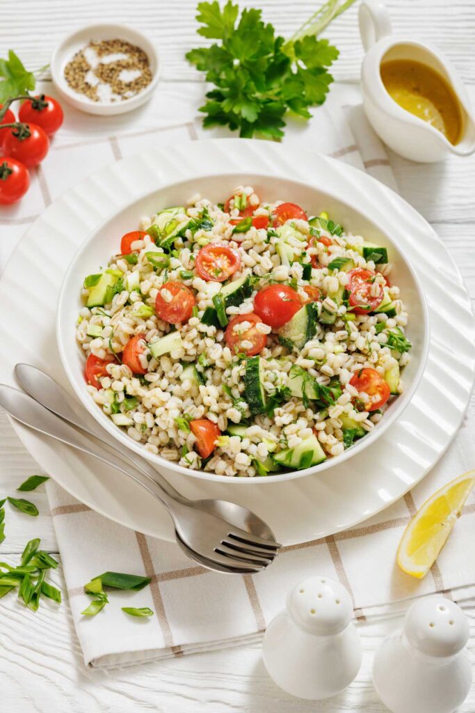 A bowl of barley salad with cherry tomatoes, cucumber, and herbs, served with a fork and spoon on a plate, surrounded by dressing, parsley, lemon, and condiments.