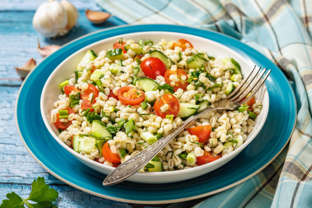 A bowl of barley salad with cherry tomatoes, cucumber, parsley, and green onions, served with a fork on a blue plate. Garlic bulbs and a checked napkin are in the background.