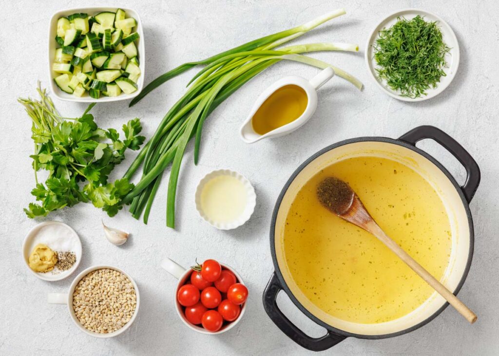 A pot of yellow soup with a wooden spoon is surrounded by fresh herbs, chopped vegetables, cherry tomatoes, seasonings, grains, and a small pitcher of olive oil on a white surface.