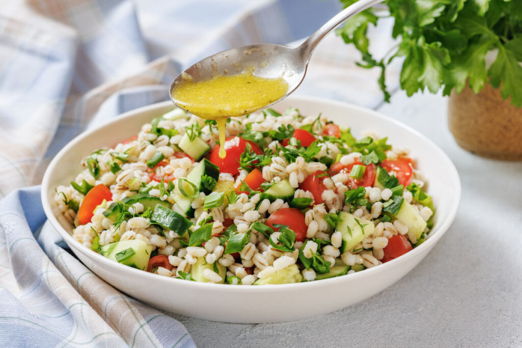 A spoon drizzles dressing over a bowl of barley salad with chopped tomatoes, cucumbers, green onions, and herbs on a light table.