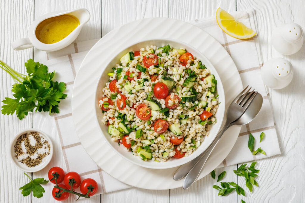 A bowl of barley salad with cherry tomatoes, cucumber, parsley, and green onions, served with a lemon wedge, salad dressing, fresh herbs, and utensils on a white table.