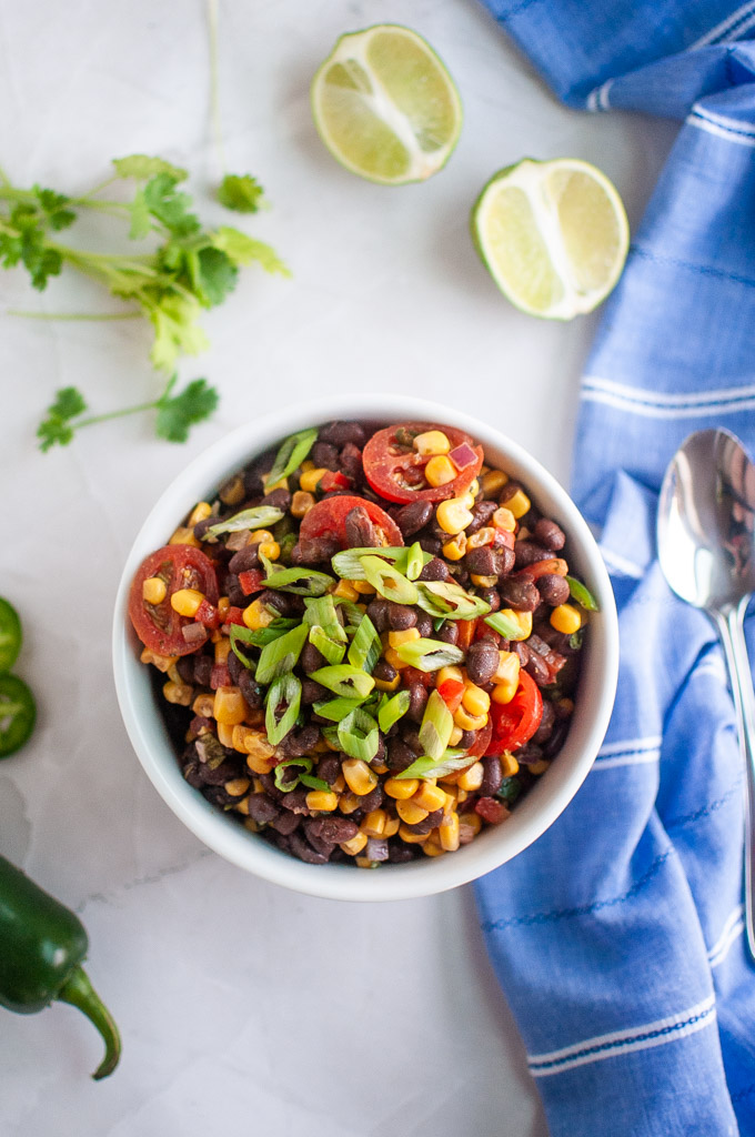 A bowl of black bean and corn salad with cherry tomatoes and chopped green onions, surrounded by lime halves, cilantro, a jalapeño, and a blue napkin with a spoon.