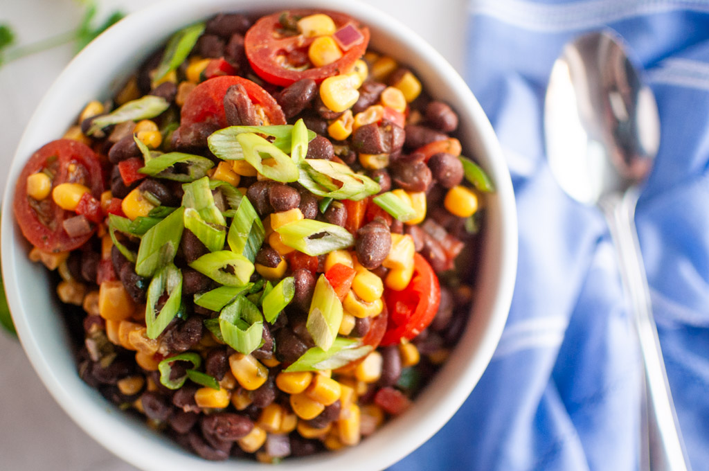 A white bowl filled with Black Bean-Corn Crunch Salad&mdash;featuring black beans, corn, cherry tomatoes, and chopped green onions&mdash;sits next to a blue napkin and a spoon.
