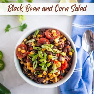 A bowl of black bean and corn salad with tomatoes and chopped green onions, placed on a white surface next to a blue napkin and spoon.