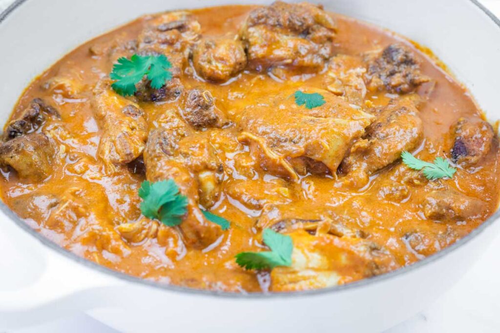 A white pot filled with chicken pieces in a thick, orange-colored curry sauce, garnished with a few cilantro leaves.