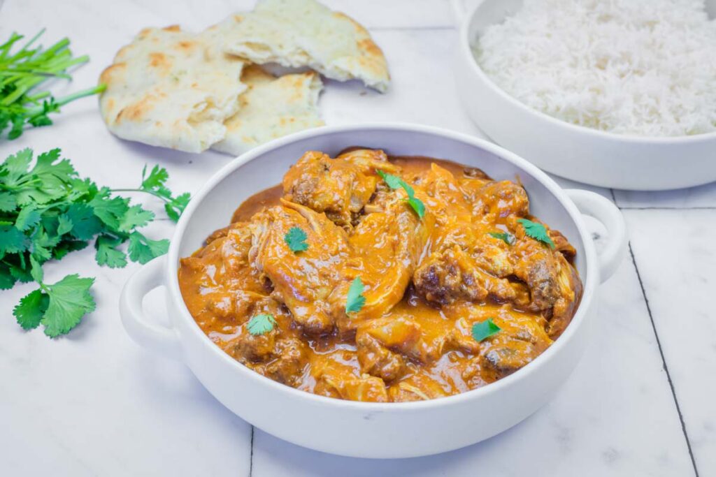 A bowl of chicken curry garnished with cilantro, served with naan bread and a bowl of white rice on a marble surface.