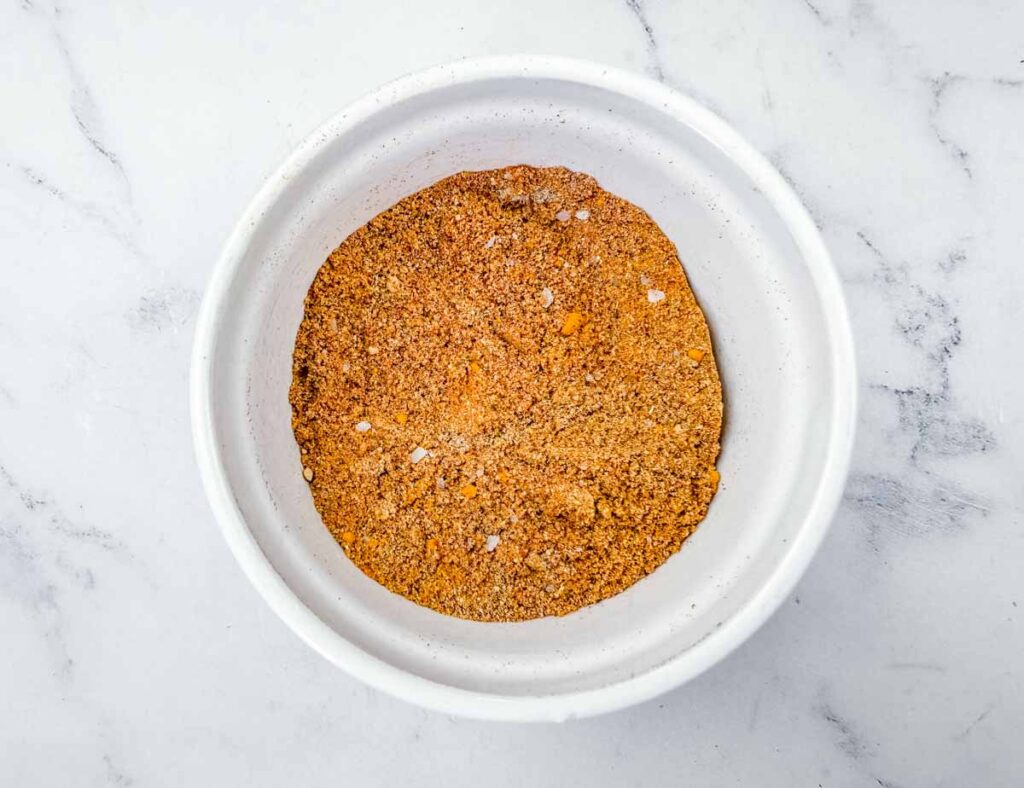 A bowl filled with a reddish-brown dry spice blend sits on a white marble surface.