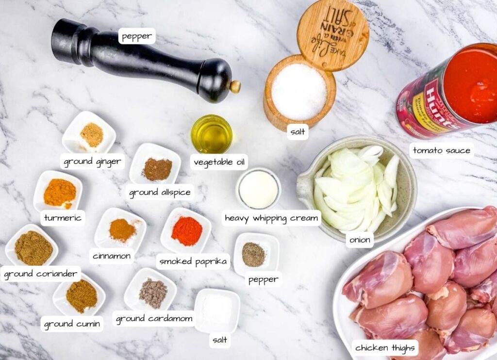 Ingredients for a recipe laid out on a marble surface, including spices, vegetable oil, salt, pepper, tomato sauce, onion slices, heavy whipping cream, and raw chicken thighs.