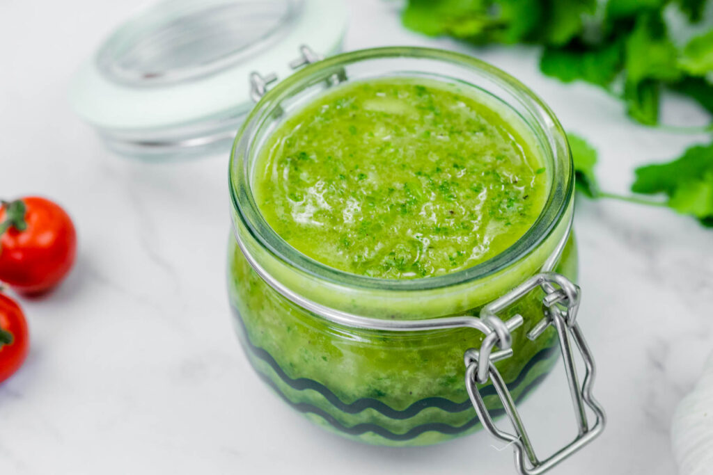 A glass jar filled with green sauce or salsa sits on a white surface next to fresh cilantro and cherry tomatoes.