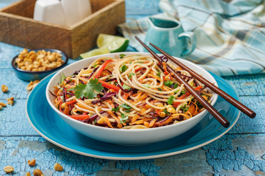 A bowl of colorful noodle salad with vegetables, topped with cilantro, placed on a blue plate with chopsticks, with peanuts and lime on the side.