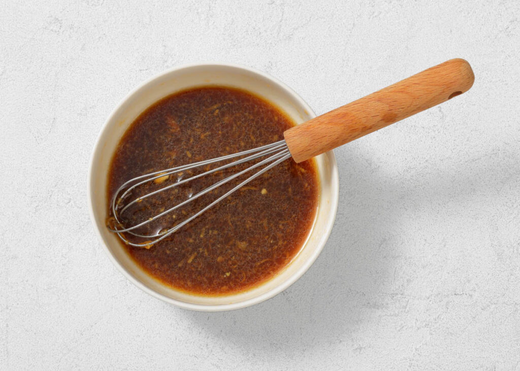A bowl of dark brown sauce with a metal whisk and wooden handle on a light textured surface.