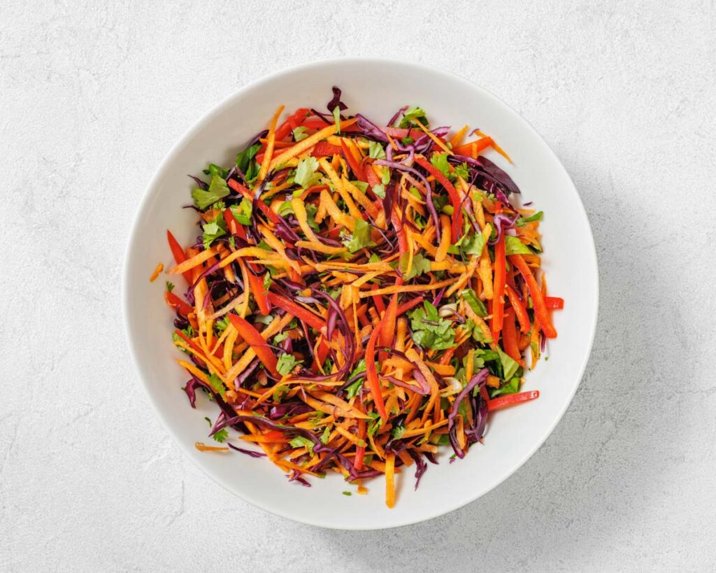 A white bowl filled with a colorful salad made of shredded carrots, red cabbage, red bell pepper, and chopped cilantro on a light surface.