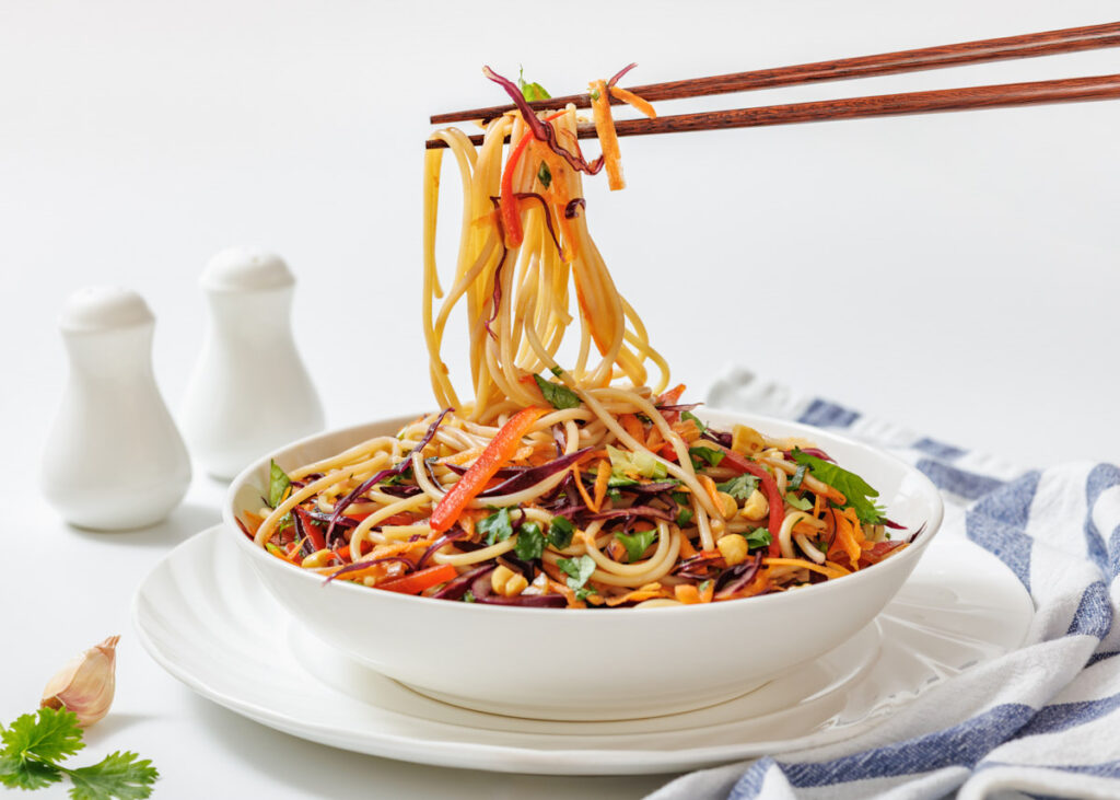 A bowl of vegetable noodle salad with chopsticks lifting a portion, next to salt and pepper shakers on a white surface.