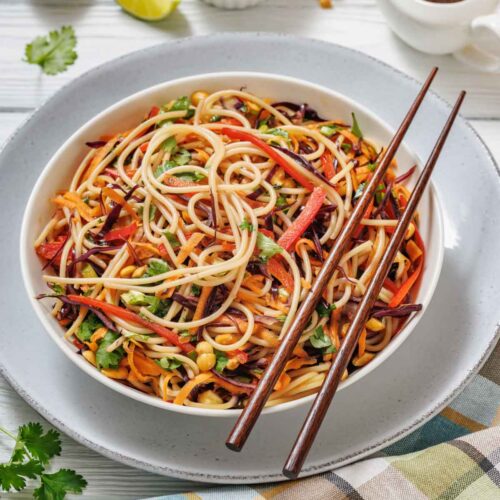 A bowl of noodle salad with vegetables and peanuts, served with chopsticks. Lime wedges, peanuts, and a small jug of sauce are nearby on a white table.