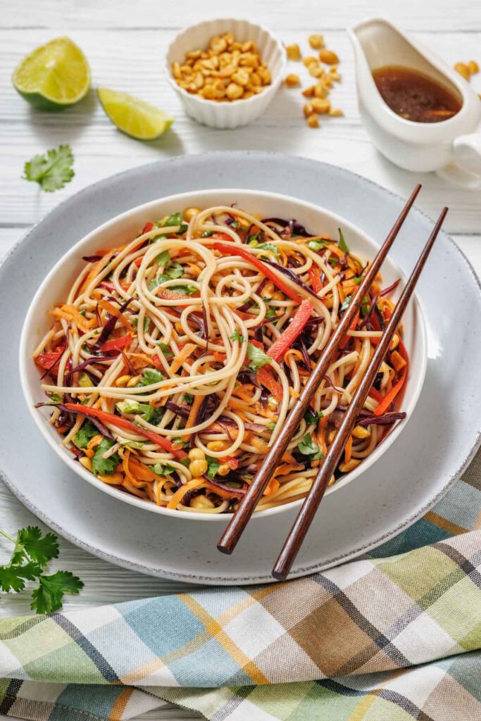 A bowl of noodle salad with vegetables and peanuts, served with chopsticks. Lime wedges, peanuts, and a small jug of sauce are nearby on a white table.