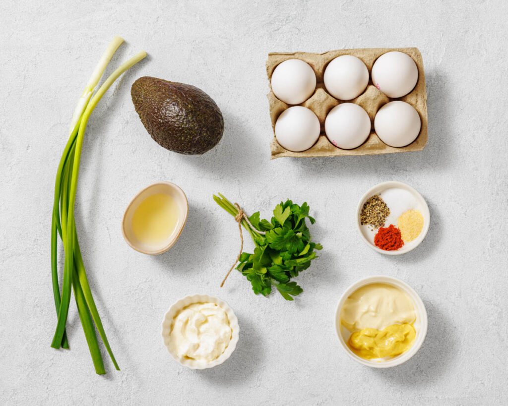 Overhead shot of avocado egg salad ingredients, including eggs, avocado, herbs, mayonnaise, mustard, and spices, arranged on a light surface.