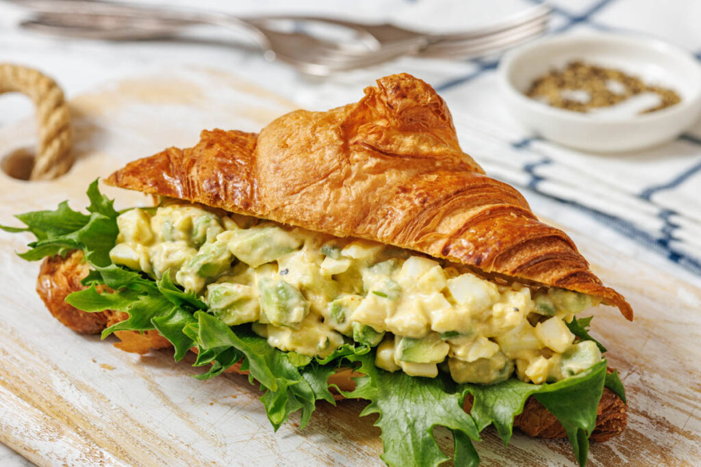 A close-up shot of an avocado egg salad croissant sandwich with lettuce on a light wooden cutting board.