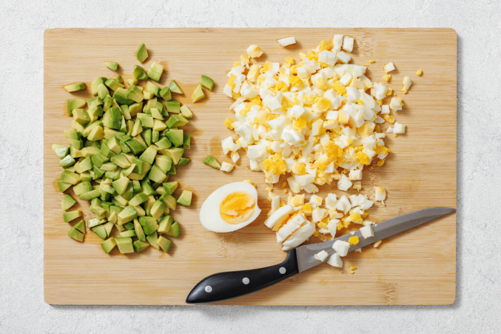 Overhead shot of chopped avocado and eggs on a light wooden cutting board, with a knife.