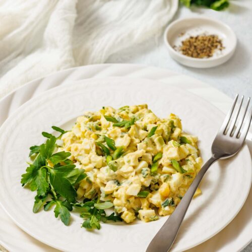A white plate with avocado egg salad, garnished with herbs, is shown with a fork, cloth, and a small bowl of spices.