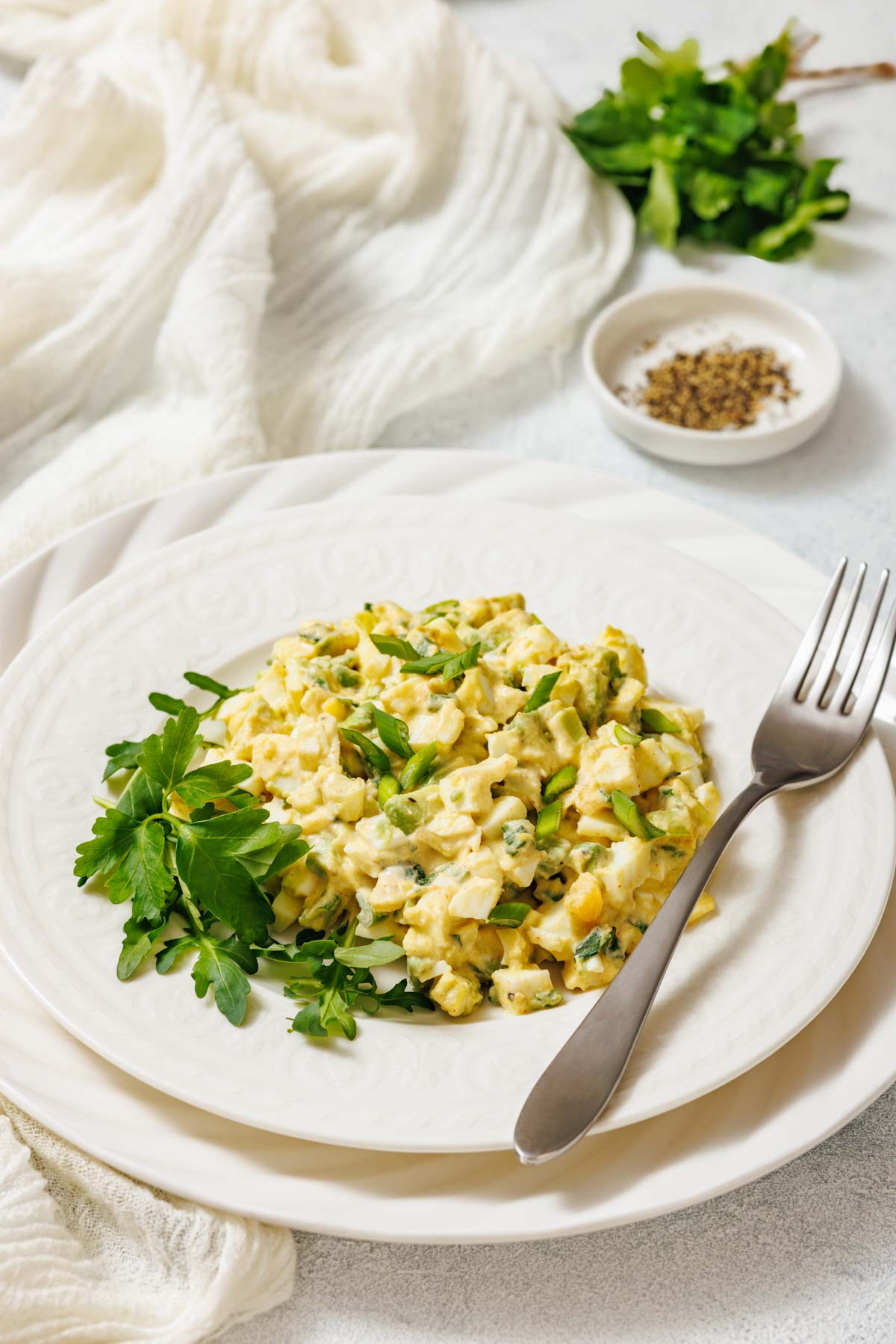 A white plate with avocado egg salad, garnished with herbs, is shown with a fork, cloth, and a small bowl of spices.