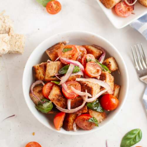 Overhead shot of panzanella salad in a white bowl, featuring toasted bread cubes, cherry tomatoes, red onion, and basil leaves, with a fork and cloth.