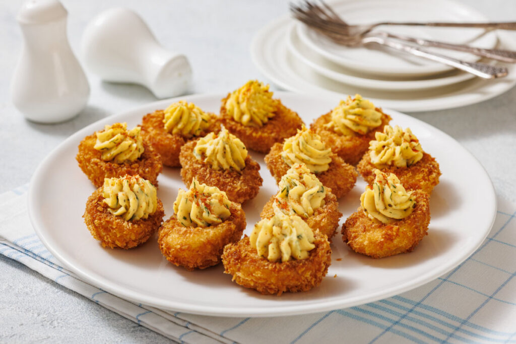 A white plate with ten fried deviled eggs, garnished with herbs, is shown with salt and pepper shakers, plates, forks, and a cloth in the background.
