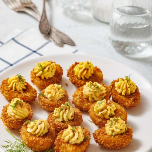 A white plate with ten fried deviled eggs, garnished with dill, is shown with forks, glasses, and a cloth in the background.
