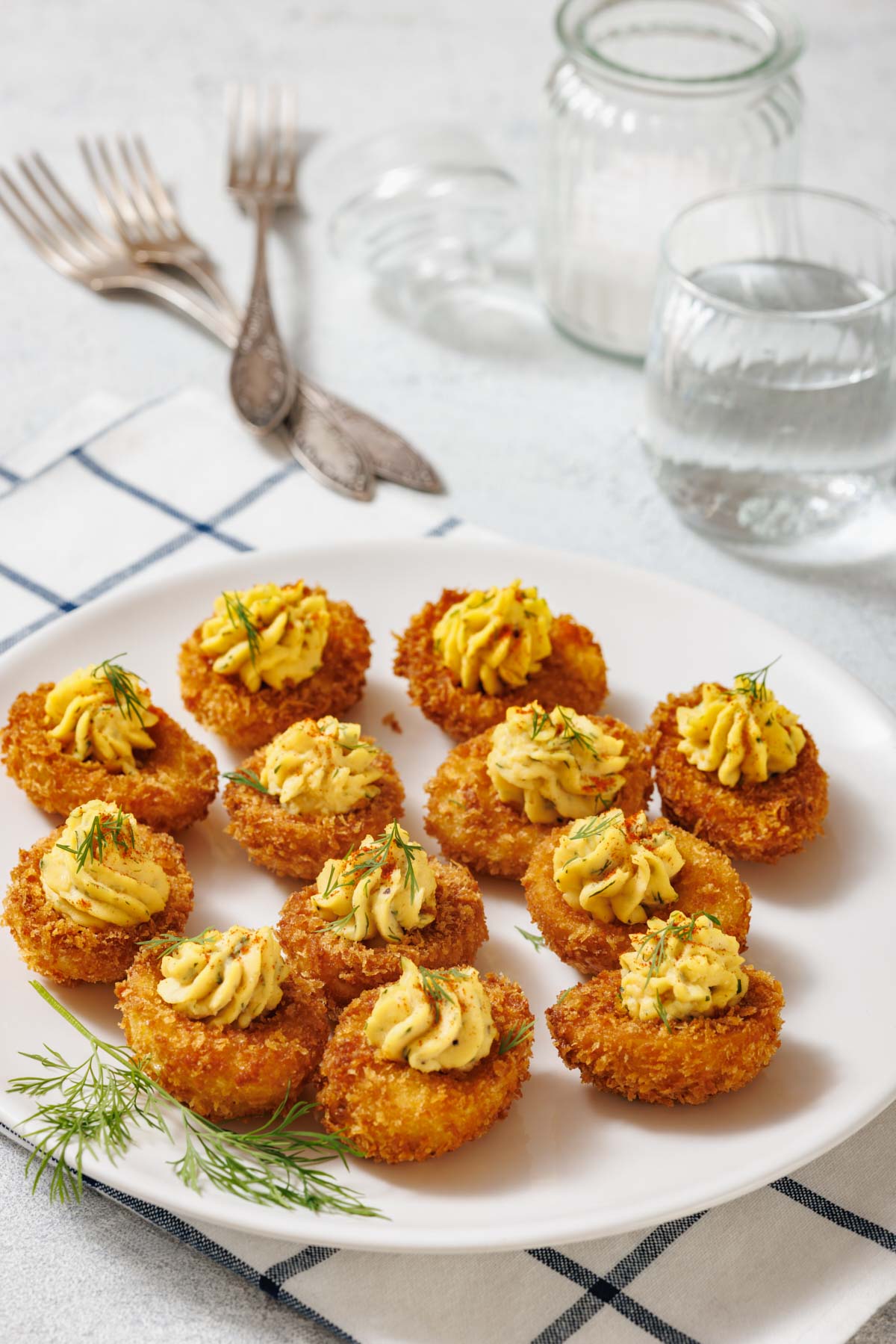 A white plate with ten fried deviled eggs, garnished with dill, is shown with forks, glasses, and a cloth in the background.