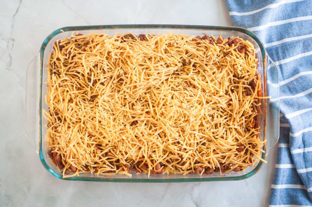 Overhead shot of a casserole with beans, ground beef, and shredded cheese in a clear glass baking dish, with a blue striped cloth beside it.