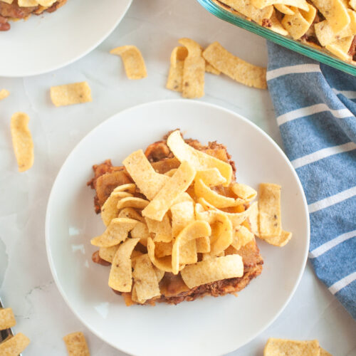 Overhead view of a serving of Frito pie topped with crunchy corn chips on a white plate, surrounded by scattered chips and a baking dish filled with the same dish nearby.