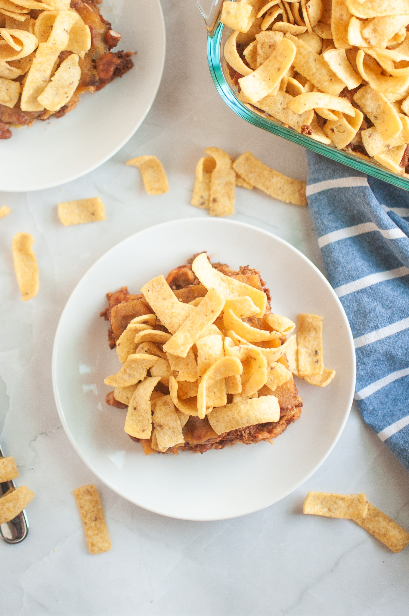 Overhead view of a serving of Frito pie topped with crunchy corn chips on a white plate, surrounded by scattered chips and a baking dish filled with the same dish nearby.