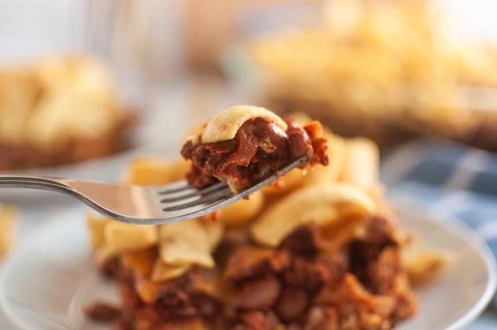 Close-up of a fork holding a bite of Frito pie, showing layers of seasoned beans, chili, and crunchy corn chips with a blurred plate of the dish in the background.