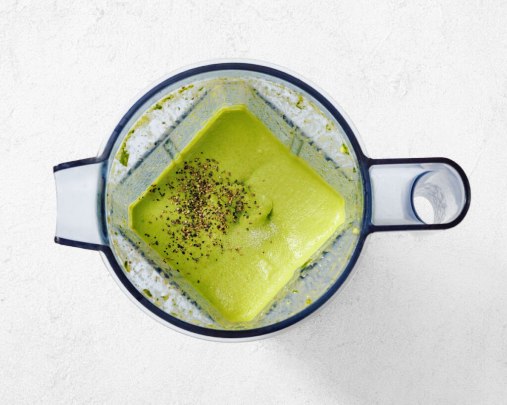 A blender filled with green sauce topped with black pepper sits on a white surface.
