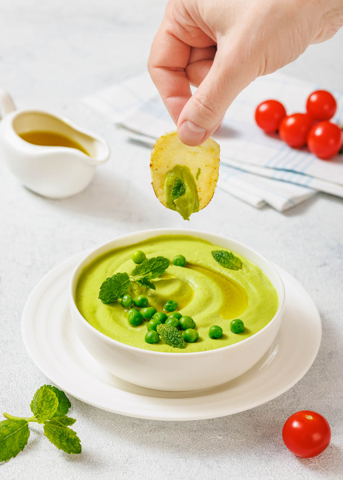 A hand dips a chip into a bowl of green pea hummus, garnished with peas and mint.