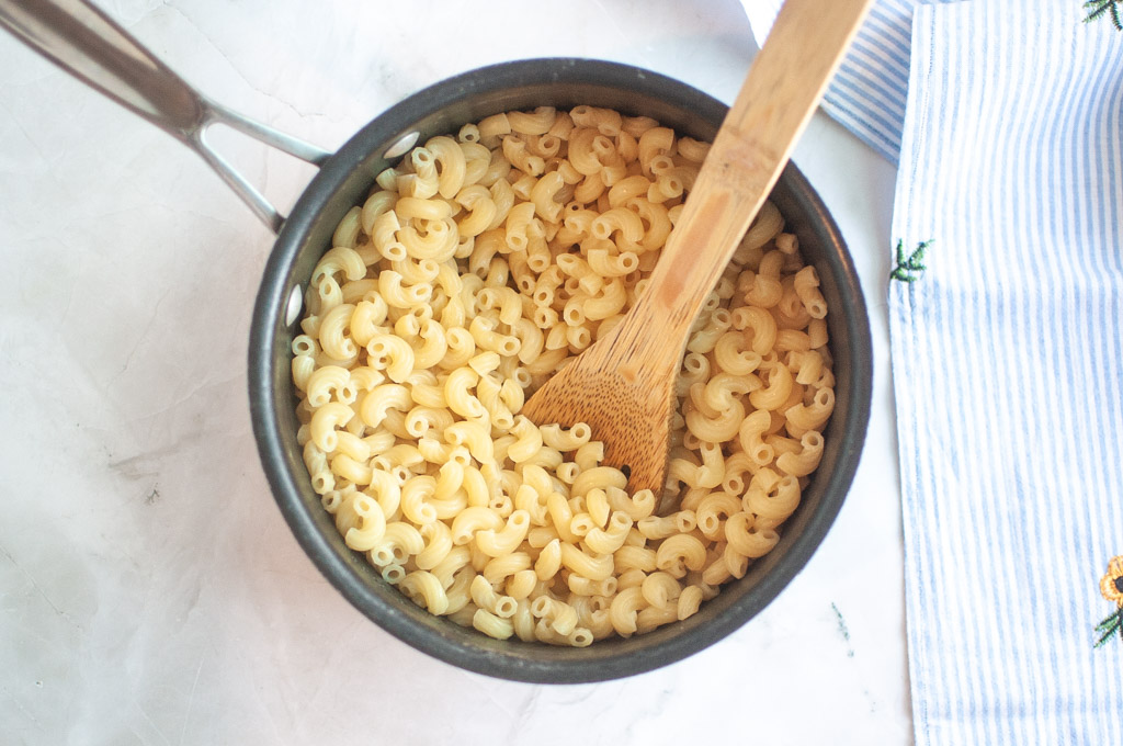 Overhead shot of cooked elbow macaroni in a dark pot with a wooden spoon, on a light surface with a striped cloth.