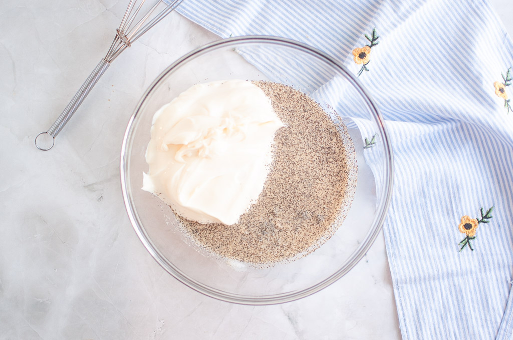Overhead shot of mayonnaise and seasonings in a glass bowl, with a whisk and striped cloth nearby.