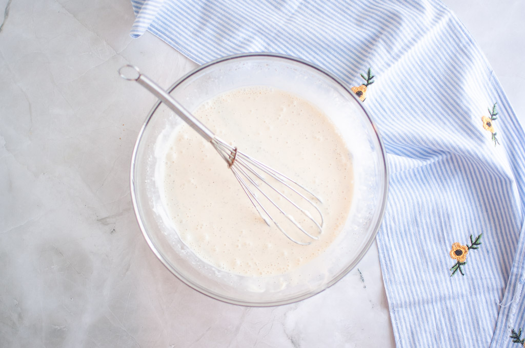 Overhead shot of a whisked dressing in a glass bowl, with a whisk inside and a striped cloth nearby.