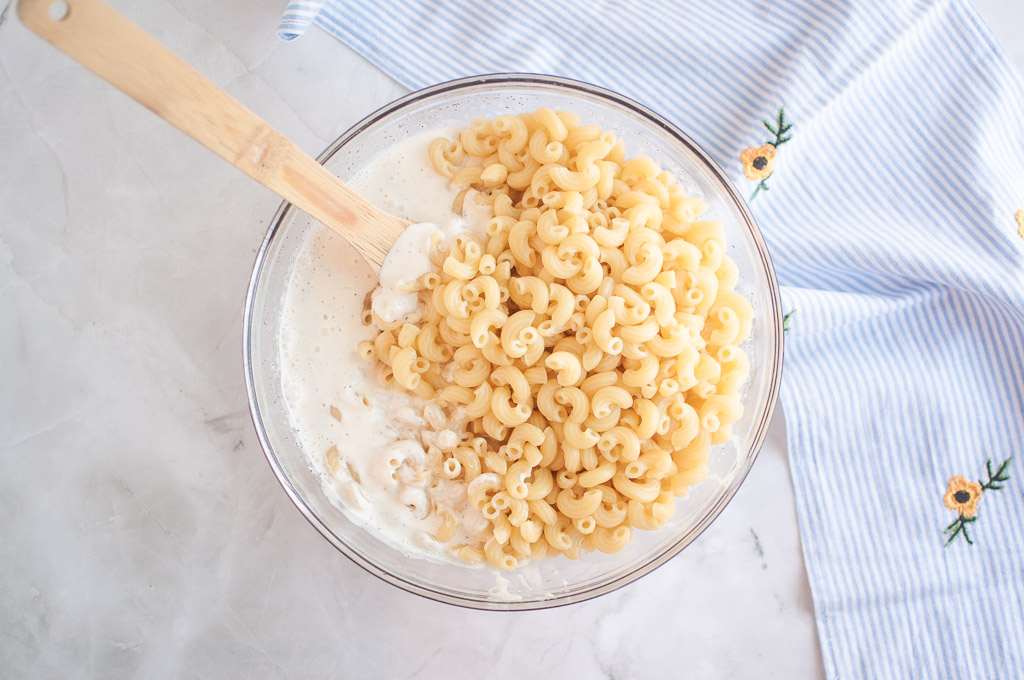 Overhead shot of cooked macaroni being added to dressing in a glass bowl, with a wooden spoon and striped cloth nearby.