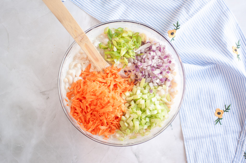 Overhead shot of macaroni salad with added vegetables in a glass bowl, with a wooden spoon and striped cloth nearby.