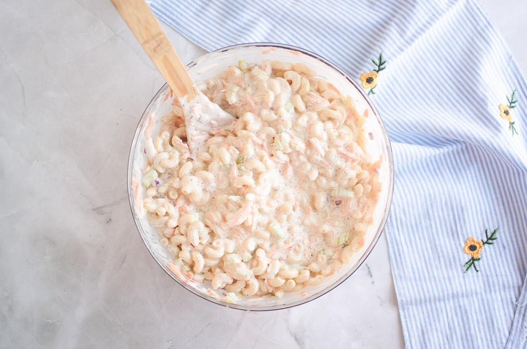 Overhead shot of macaroni salad in a glass bowl, being mixed with a wooden spoon, with a striped cloth nearby.