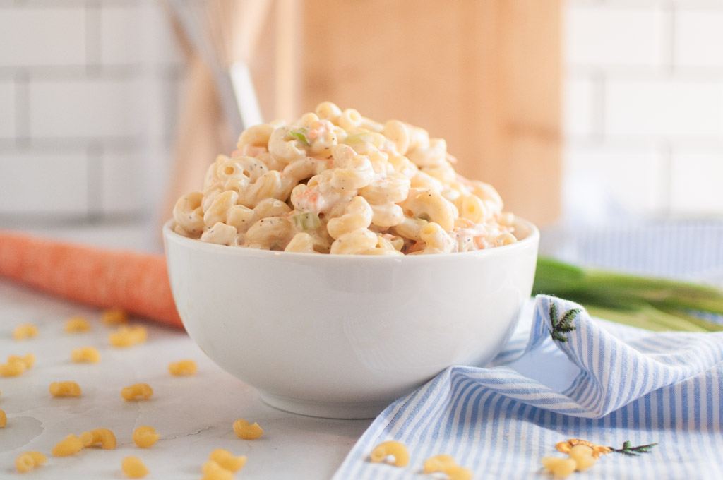 A white bowl filled with Hawaiian macaroni salad, containing elbow macaroni and a creamy dressing, is shown with carrots and scallions on the side.