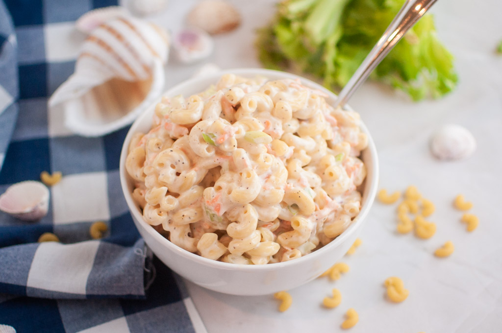 A white bowl filled with macaroni salad, containing elbow macaroni and a creamy dressing, is shown with a spoon inside. Garlic cloves and lettuce are visible around the bowl.