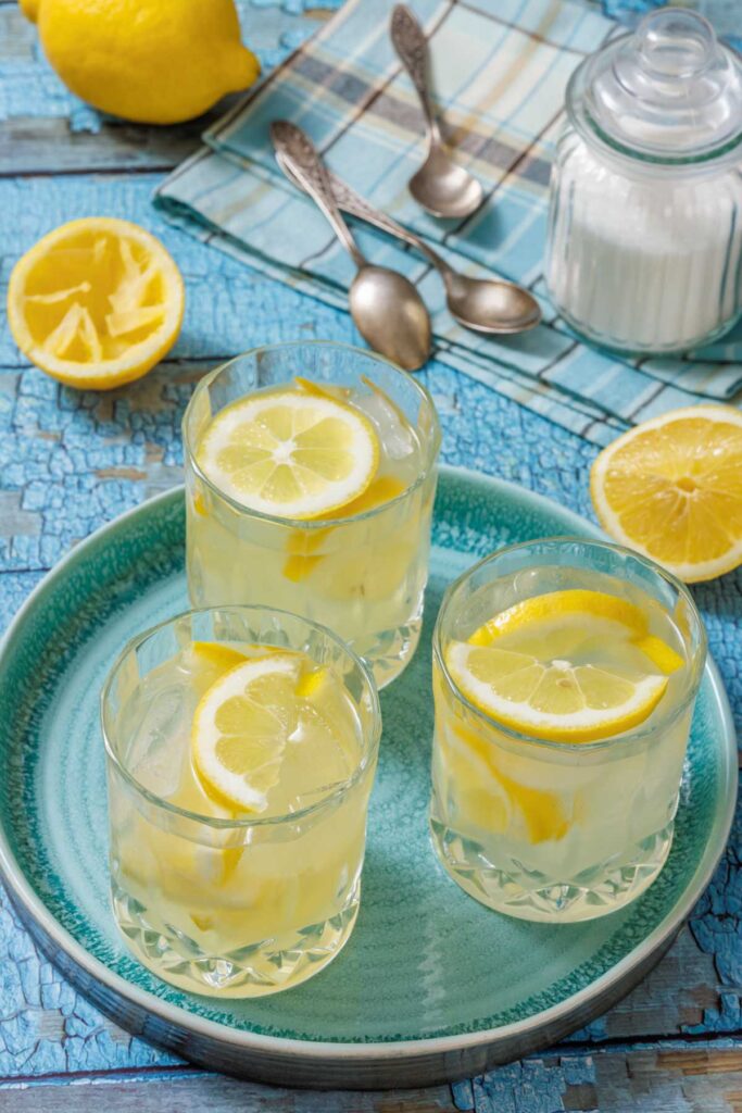 Three glasses of lemonade with lemon slices on a blue plate, placed on a distressed blue wooden table. Two silver spoons, a glass jar of sugar, and additional lemon halves are arranged around the plate.