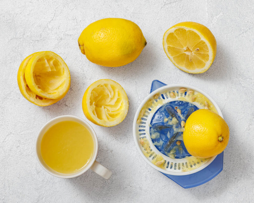 Flat lay of freshly squeezed lemons, a manual citrus juicer with pulp and seeds, and a cup filled with lemon juice on a textured light-colored surface.