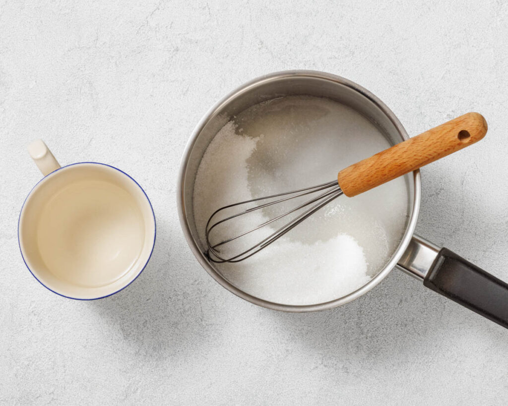 Overhead shot of a silver metal saucepan containing white granulated sugar with a wire whisk resting inside, positioned on a light gray textured background. To the left is a small, white cup with a blue rim, filled with a clear liquid. The saucepan has a black handle and the whisk has a wooden handle. The surface is a light gray with a subtle speckled pattern.